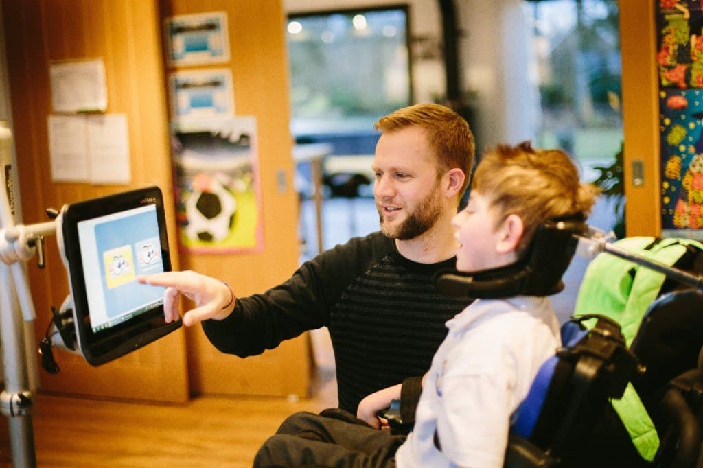 A man points to pictures on a screen mounted beside a boy in a wheelchair.