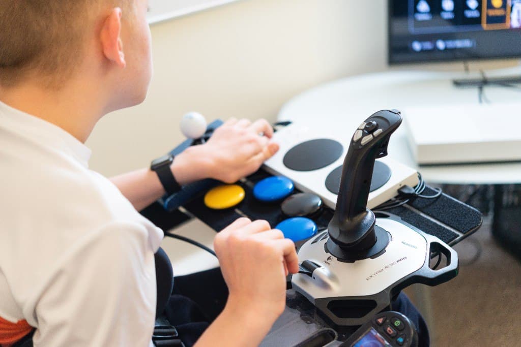 A person uses a joystick and large coloured buttons on a tray, with a screen in front of them.