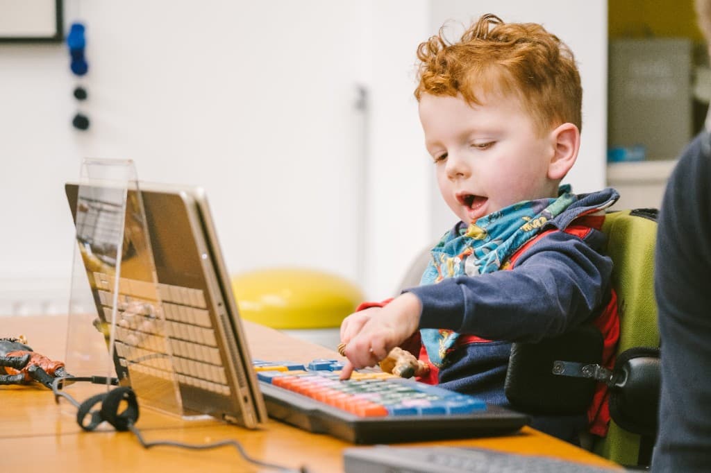 A young boy in a supportive chair presses a colourful keyboard while looking at a laptop.