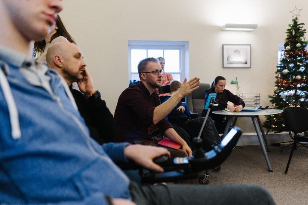 People seated together in a meeting, while a man in a wheelchair raises his hand as he speaks.