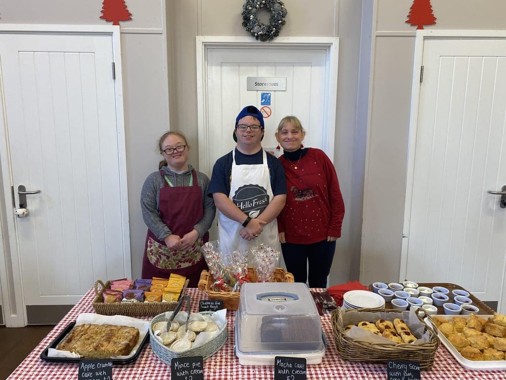 Three people stand behind a table of cakes, mince pies and scones, with Christmas decorations on the wall behind them.